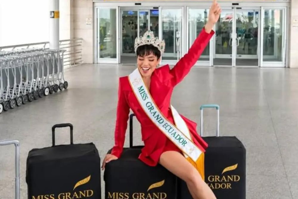 Mujer con corona y banda Miss Grand Ecuador saluda sonriente sentada sobre maletas en un aeropuerto.