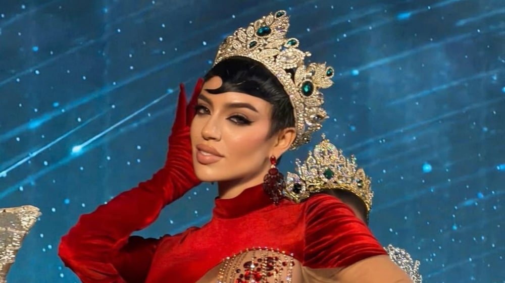 Mujer con corona de reina cubierta de piedras verdes, vestido rojo y guantes largos, posando ante un fondo azul con estrellas.