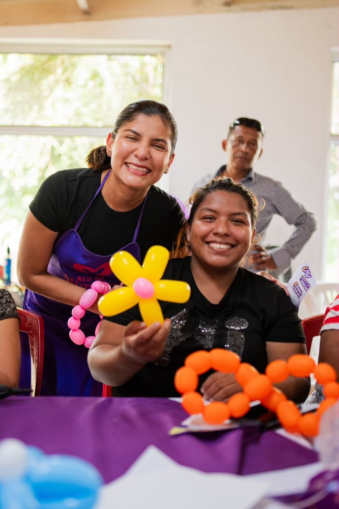 Dos mujeres sonríen y muestran una flor hecha con globos, sentadas a una mesa con accesorios de globos de colores.