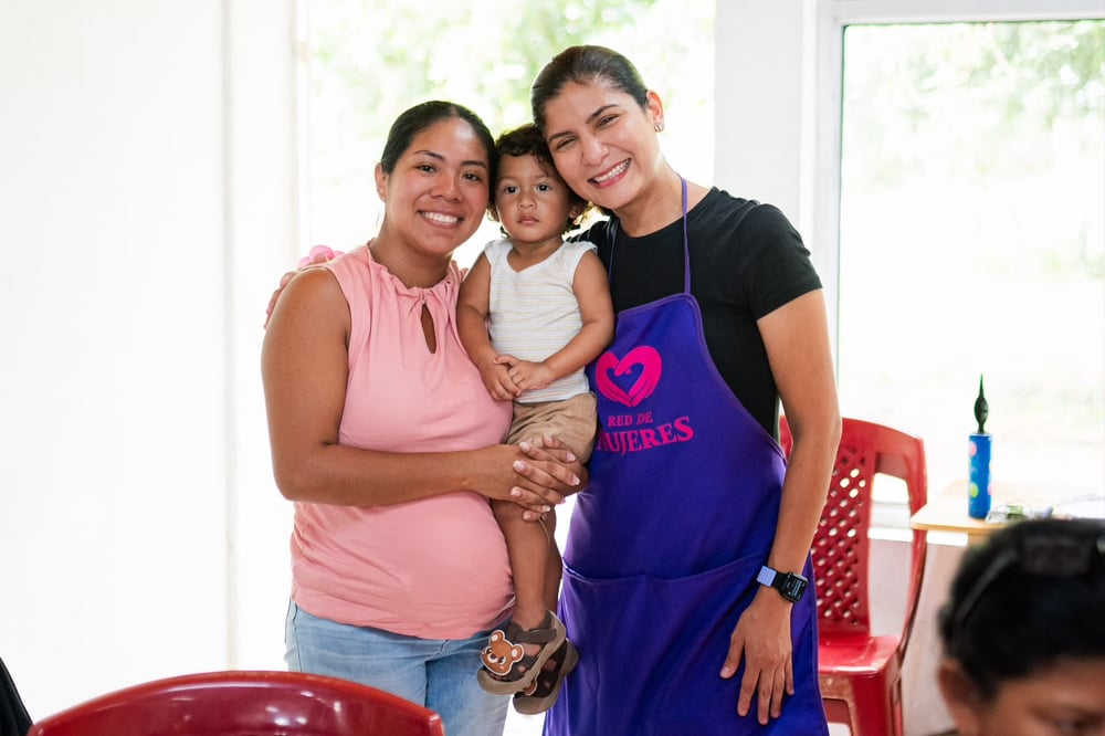 Dos mujeres sonrientes con un niño en brazos, una usa delantal morado con el logo Red de Mujeres.