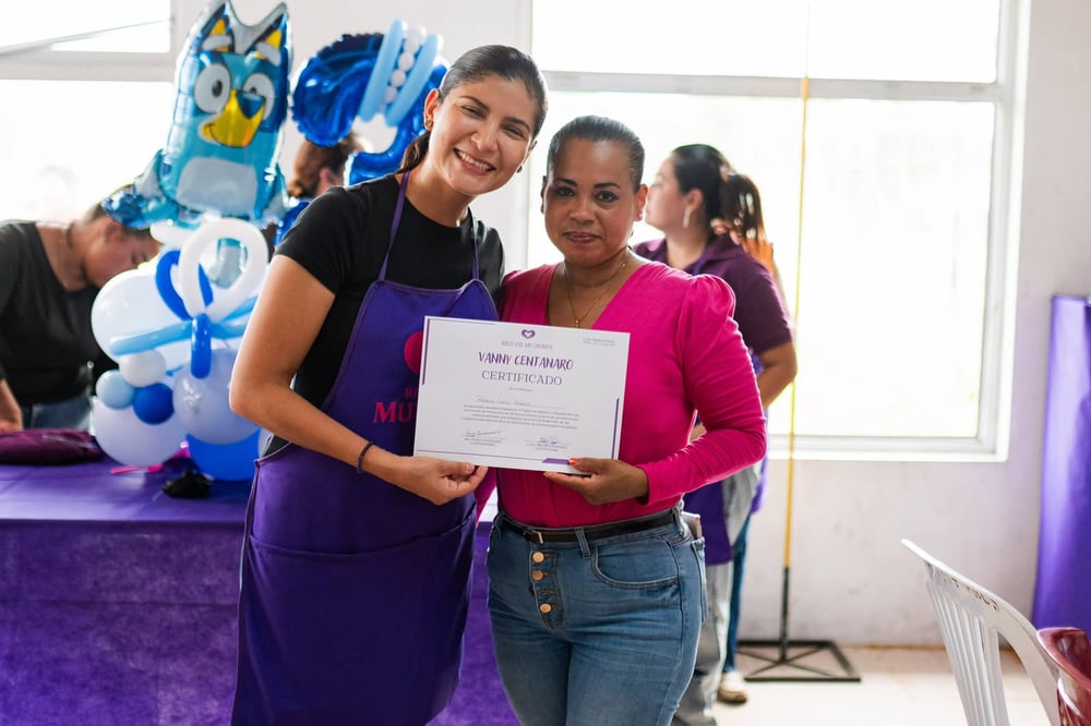 Dos mujeres sonrientes sostienen un certificado de la Red de Mujeres junto a globos, en un taller de capacitación.