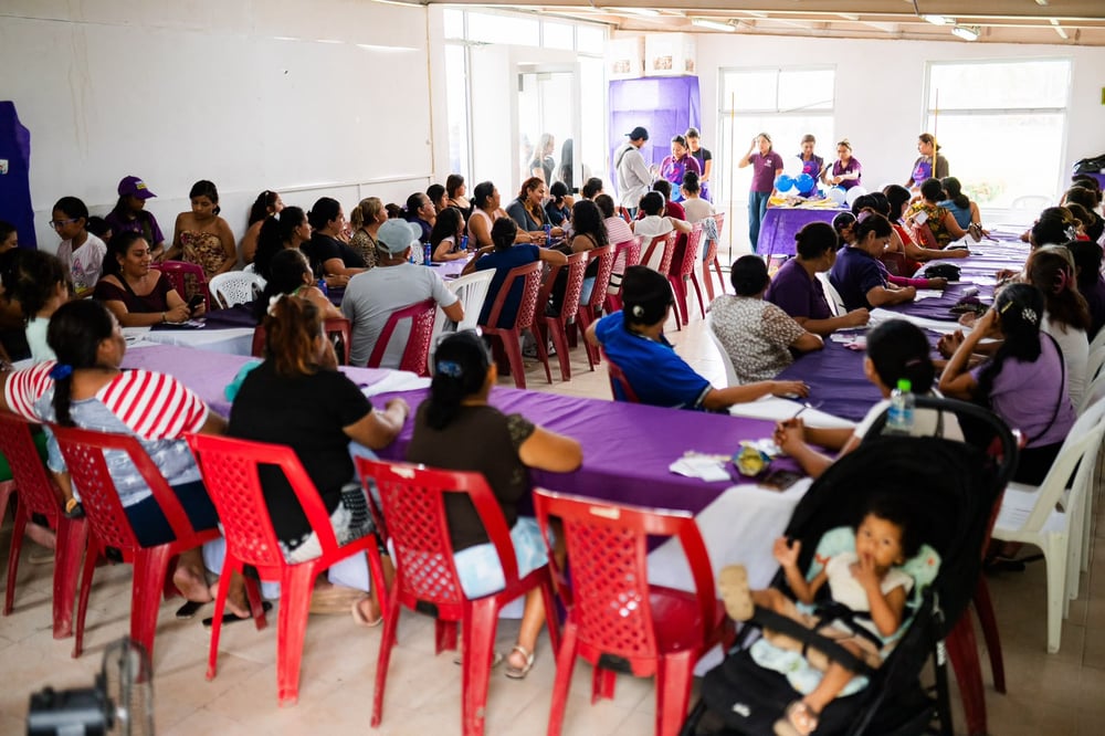 Mujeres sentadas en mesas con manteles morados durante un taller en un salón, con un bebé en coche al frente.