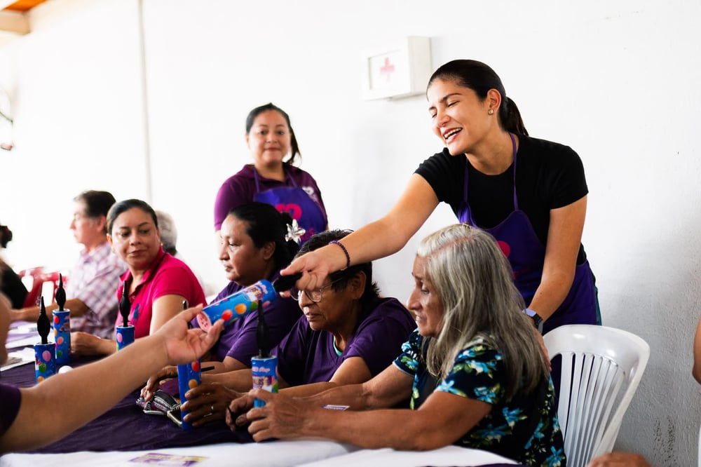 Mujeres con delantal morado sentadas a la mesa reciben serpentinas, mientras una organizadora les reparte entre risas.