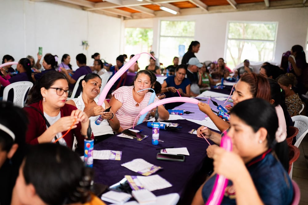 Mujeres sentadas en mesas largas inflando globos para manualidades durante un taller en un salón techado.