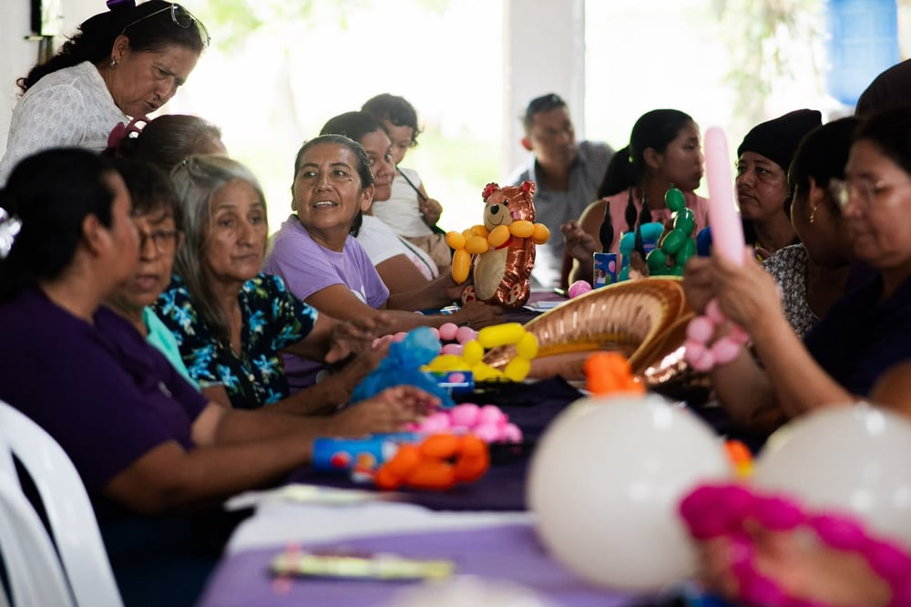Mujeres sentadas alrededor de una mesa hacen figuras con globos de colores en un taller bajo techo.