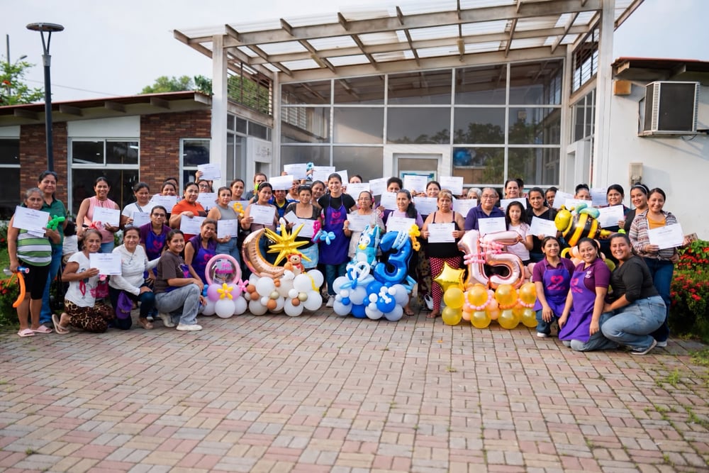 Grupo numeroso de mujeres con certificados en mano, posando junto a decoraciones de globos frente a un edificio.